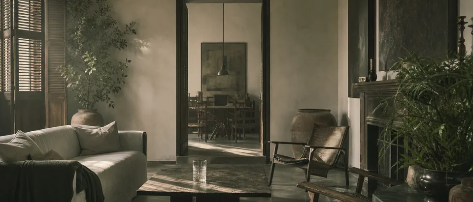 Sala de una casa completamente amueblada y sin nadie dentro, con luz fría entrando por las persianas entreabiertas y un vaso olvidado sobre la mesa baja.