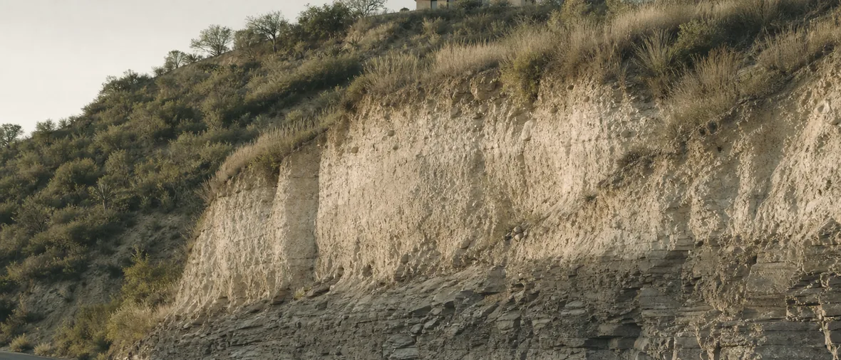 Corte de una ladera que deja ver el terreno en sección: roca compacta en la base, tierra clara y suelta encima, y una casa baja en lo alto de la loma.